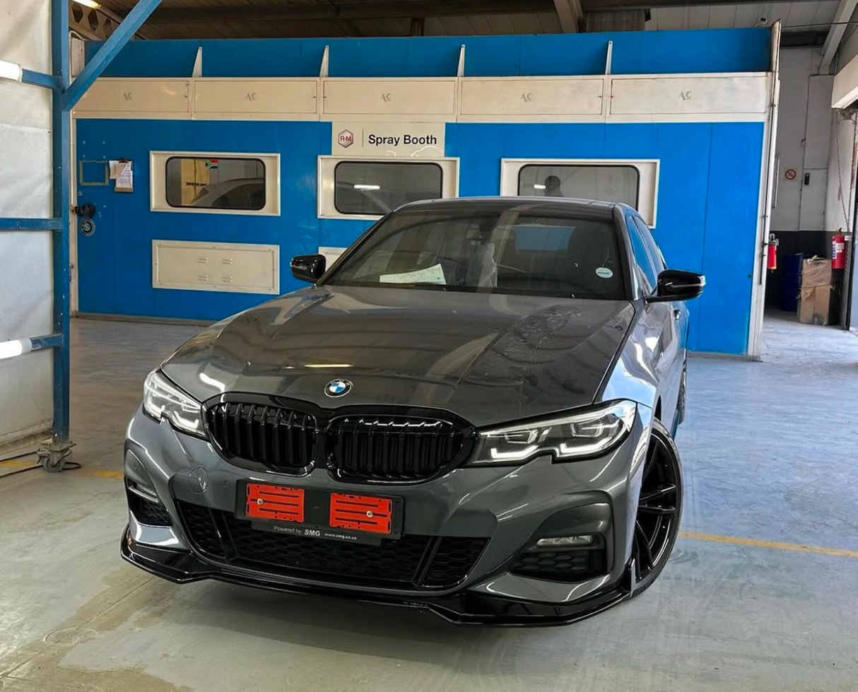 A dark grey BMW car is parked in front of a blue spray booth in a workshop. The booth includes labeled windows and doors, and the floor is concrete.