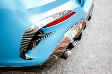 The rear section of a blue sports car with carbon fiber detailing features dual exhaust pipes, a red reflector, and is parked on a paved surface.