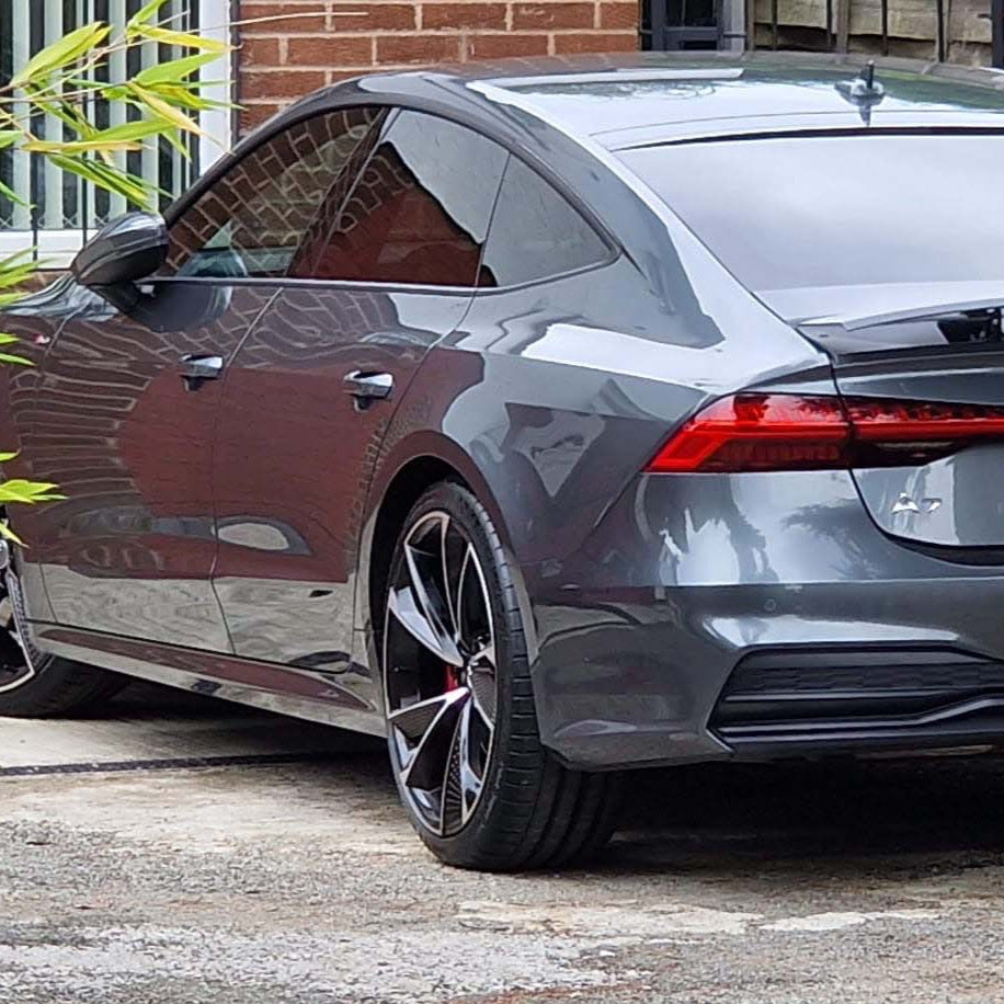 A sleek gray car is parked in a driveway next to a brick house and a wooden fence, surrounded by greenery.