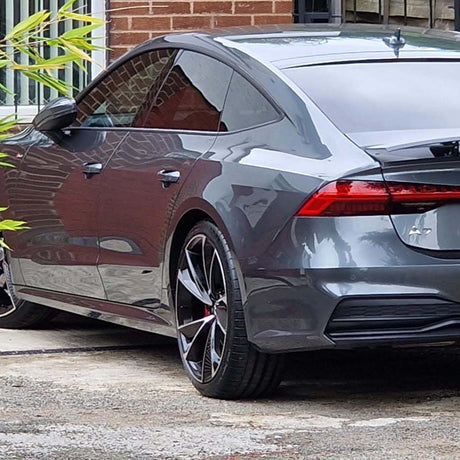 A sleek gray car is parked in a driveway next to a brick house and a wooden fence, surrounded by greenery.