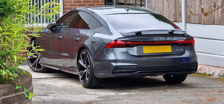 Grey Audi car parked on a driveway, facing away, next to a brick house and wooden fence, surrounded by greenery.
