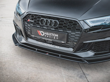 A black car's front grille displays its brand logo with a sleek design. The license plate reads "MAXTON design," and "quattro" is on the lower bumper. The car is parked on concrete.