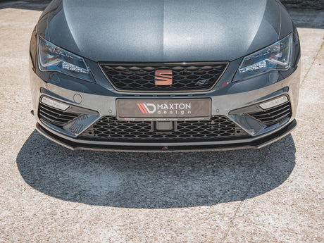 A gray car's front view features sharp headlights and a honeycomb grille. It displays a "M✪AXTON design" license plate on concrete pavement in bright sunlight.