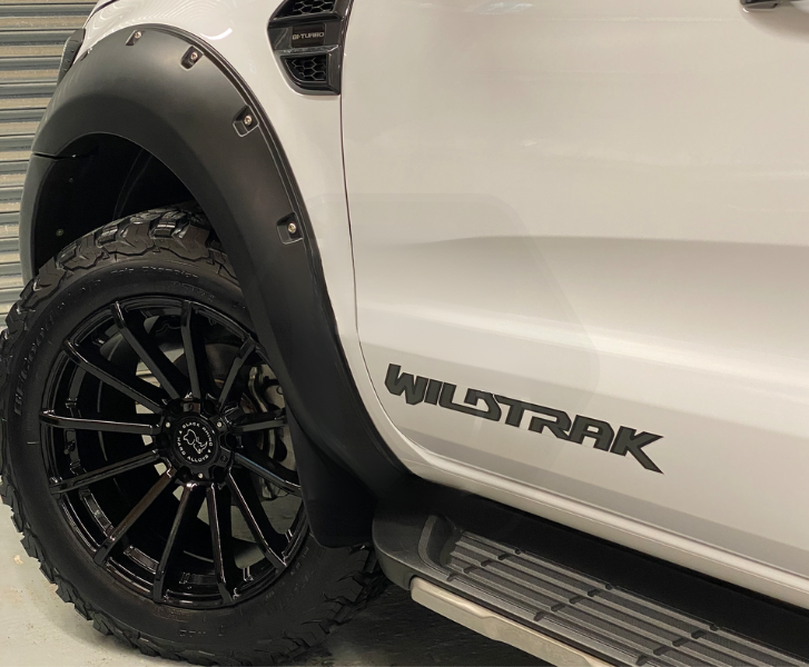 A white vehicle features black fender flares and off-road tires. Text on the side reads "WILDTRAK" and "BI-TURBO." The backdrop appears to be a garage with a gray metallic door.
