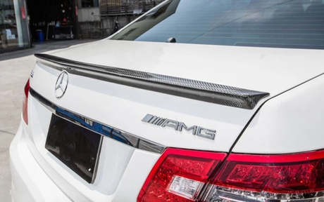 A white car with AMG and Mercedes logos features a carbon fiber spoiler on the trunk lid, parked near a building with various objects in the background.