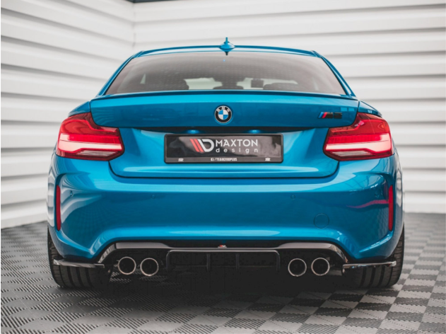 A blue BMW car is stationary, rear-facing, with dual exhausts visible. It bears the text "MAXTON design" on a plate. The background features a white, ribbed wall.
