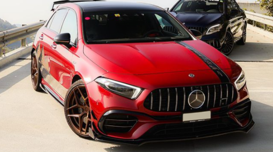 A red sports car with a black racing stripe is parked, facing forward on a sunny road. Another black car is parked behind it, both near a guardrail.