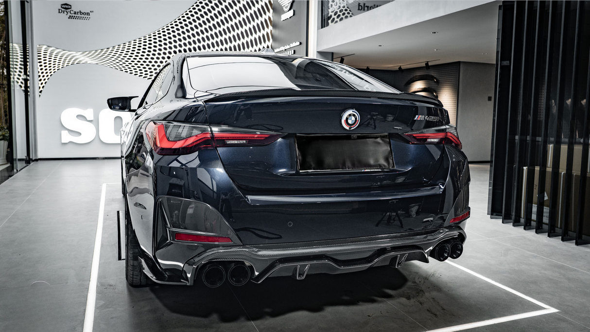A sleek, dark-colored car is parked in a showroom with a modern design. The car's rear features prominent taillights and quad exhaust pipes. The wall has text: "wrkz DryCarbon™."