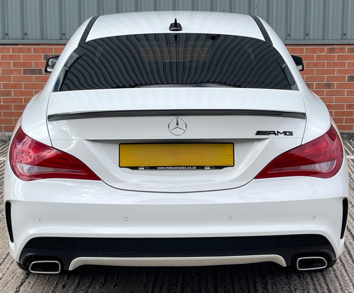 A white Mercedes-Benz AMG car is parked, featuring dual exhausts and tinted windows, against a backdrop of red brick and corrugated metal. Text: "www.mdscarsales.co.uk".