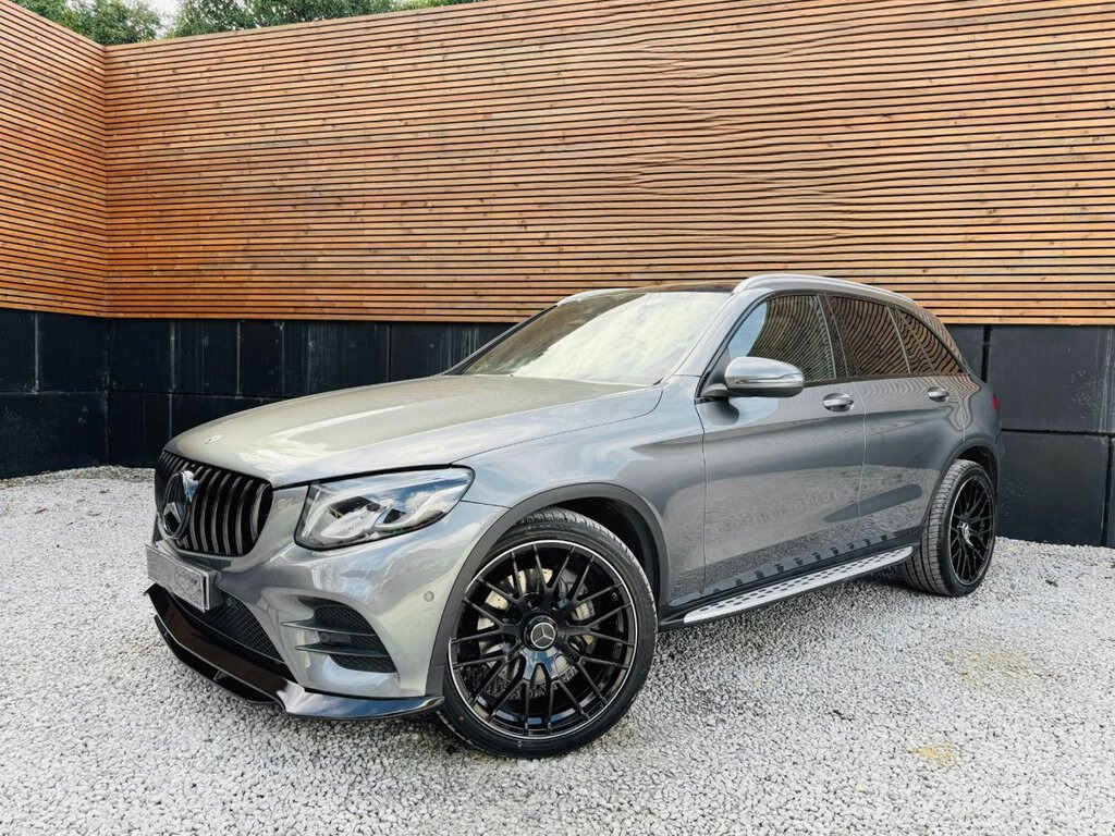 Silver SUV parked on gravel, featuring black alloy wheels and a distinctive grille. The vehicle is set against a background of horizontal wooden slats and vertical black panels.