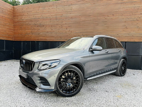 Silver SUV parked on gravel, featuring black alloy wheels and a distinctive grille. The vehicle is set against a background of horizontal wooden slats and vertical black panels.