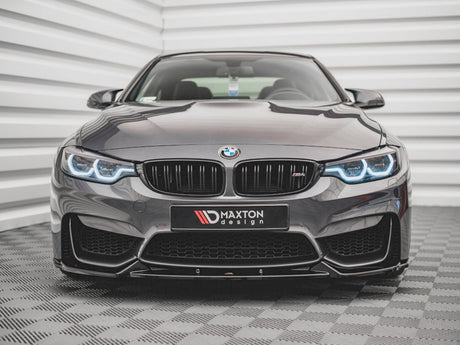 A sleek, dark BMW with illuminated headlights sits on a patterned floor, facing forward. Behind it is a textured, white wall. The front license plate reads "MAXTON design."