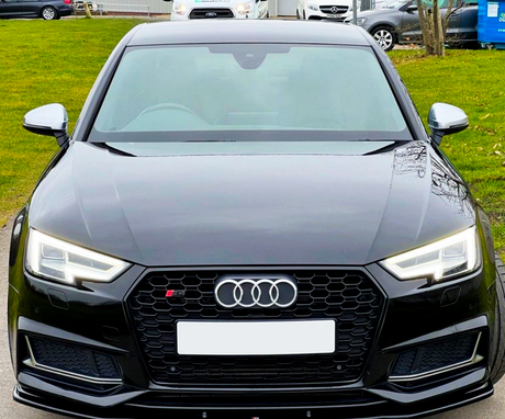 A black Audi car is parked on a road, facing forward. It features distinctive headlights and a front grille with the Audi logo. In the background, grassy areas and other parked cars are visible.