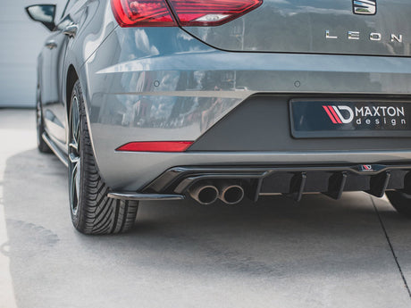 The rear of a gray car, featuring dual exhaust pipes. "LEON" and "MAXTON design" are visible. It is parked on a concrete surface, near a white building wall.