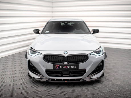 A white BMW car is stationary in a minimalist room with geometric walls and flooring. The front grille and headlights are prominent. Text on the car reads "#MAXTONIZED."