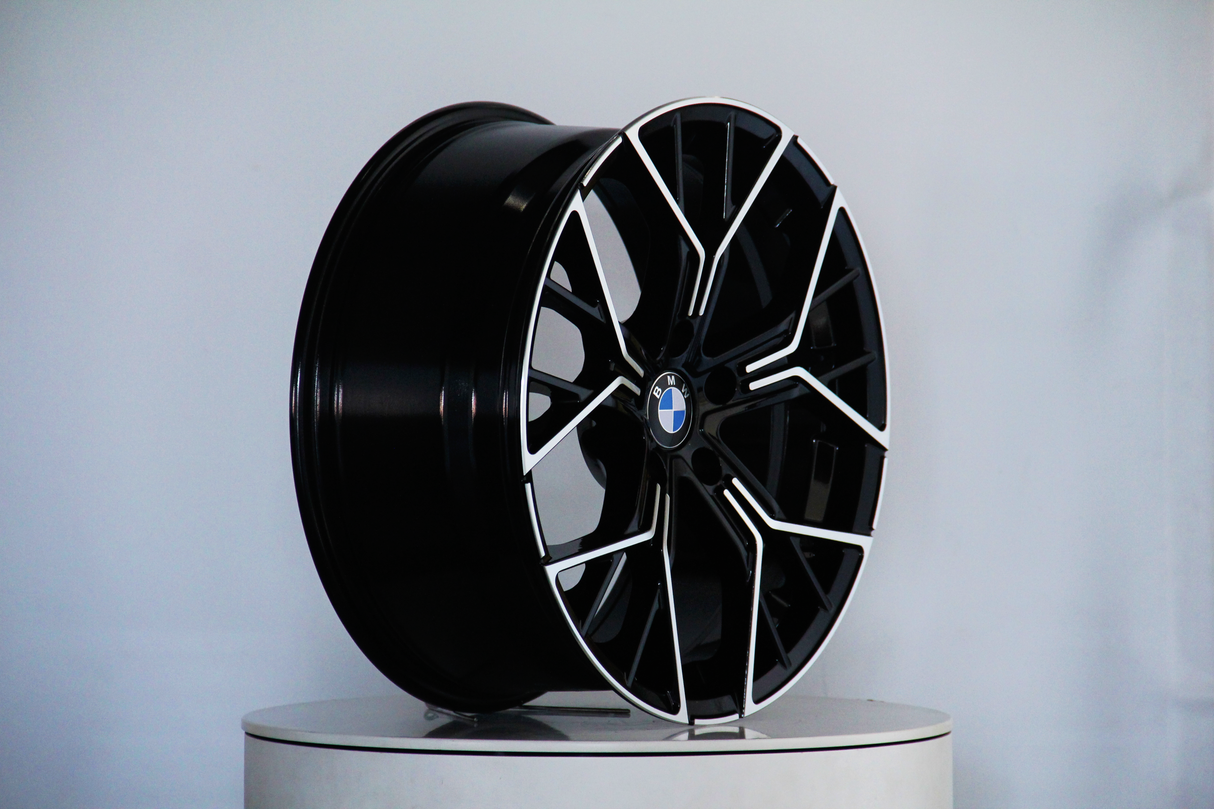 A black car wheel rim with a silver trim is displayed upright on a white surface, featuring a central blue and white BMW logo in a plain studio setting.