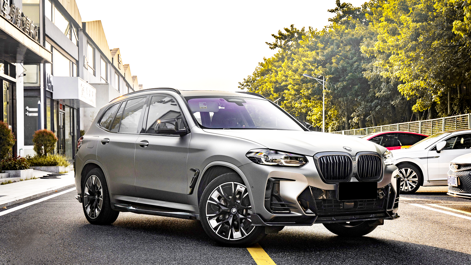 A gray SUV is parked on a sunlit street, surrounded by buildings and trees. Other cars are parked nearby.