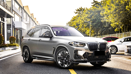 A gray SUV is parked on a sunlit street, surrounded by buildings and trees. Other cars are parked nearby.