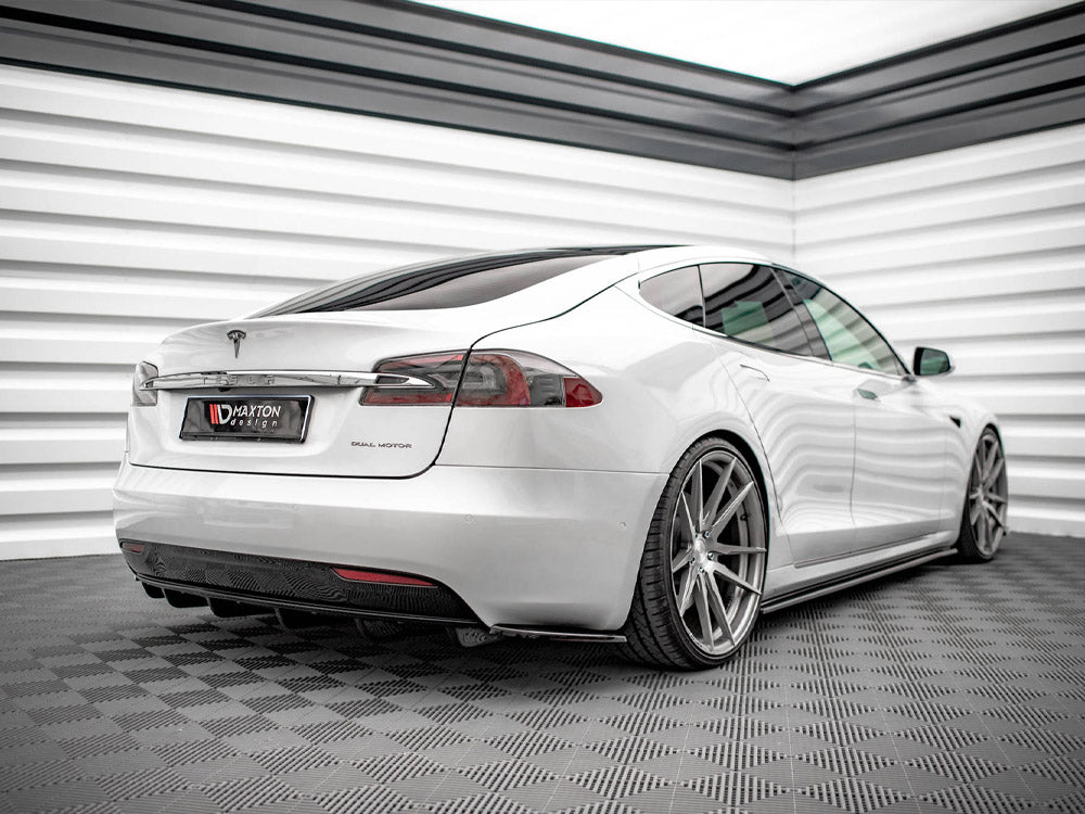 A white Tesla sedan parked in a modern showroom with checkered flooring and paneled walls. License plate reads "MAXTON design." The car has large, silver alloy wheels and tinted windows.