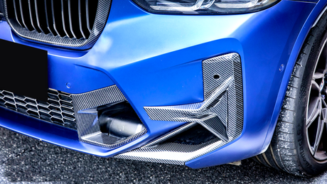 A blue car's front bumper with carbon fiber accents, features a grille and headlight, parked on a paved surface. The tire shows some white specks.
