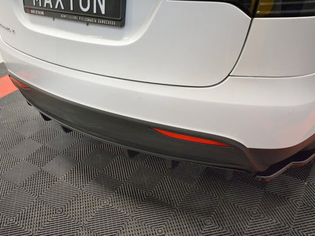 White car's rear with a black bumper and diffuser, displaying a license plate reading "MAXTON." The car is parked on a patterned floor in a showroom.