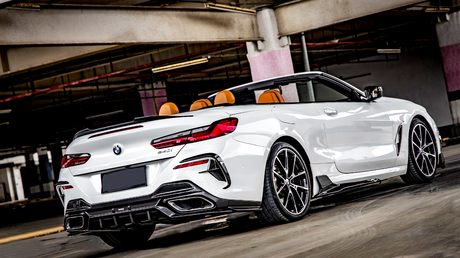 A white convertible car, with top down and orange seats visible, is parked in an empty, dimly-lit parking garage. The car has twin exhausts and modern alloy wheels.