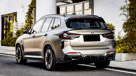 A silver SUV is parked on a city street, with surrounding greenery and modern glass buildings. The license plate and model name "iX3" are visible on the rear.