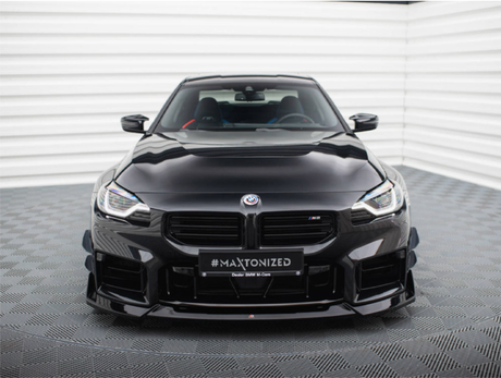 A sleek black BMW M2 faces forward, parked on a patterned floor in a bright room. The number plate reads "#MAXTONIZED."