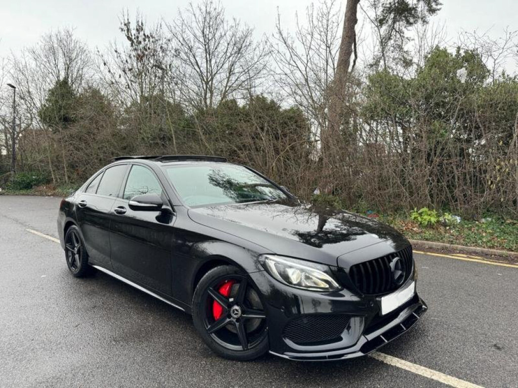 A black car with black grille and red brake calipers is parked on a wet road, surrounded by bare trees and shrubbery.