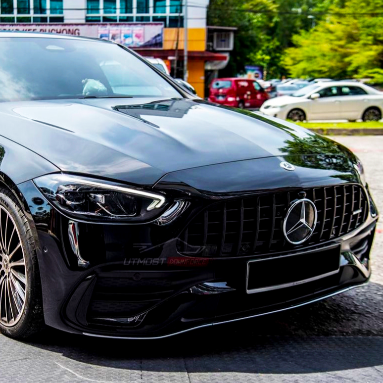 A black Mercedes-Benz car, parked on a textured surface, sits in front of a building with multiple windows. Green trees and other cars are visible in the background.