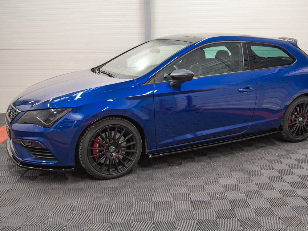A blue hatchback car is parked indoors on a textured gray floor, with black wheels and red brake calipers, surrounded by white-paneled walls.