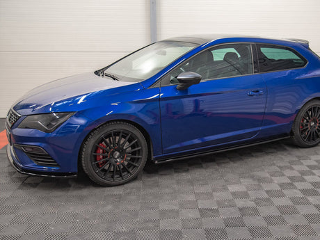 A blue hatchback car is parked indoors on a textured gray floor, with black wheels and red brake calipers, surrounded by white-paneled walls.