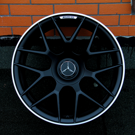 A black alloy wheel with a Mercedes-Benz logo and "AMG" branding is placed upright against an orange brick wall on a textured black surface.