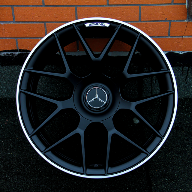 A black alloy wheel with a central Mercedes-Benz logo and AMG branding rests against an orange brick wall on a dark surface, showcasing an intricate spoke design.