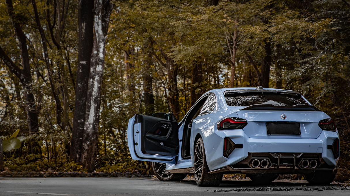 A light blue car with its driver's door open is parked on a forest road, surrounded by tall trees and dense foliage.