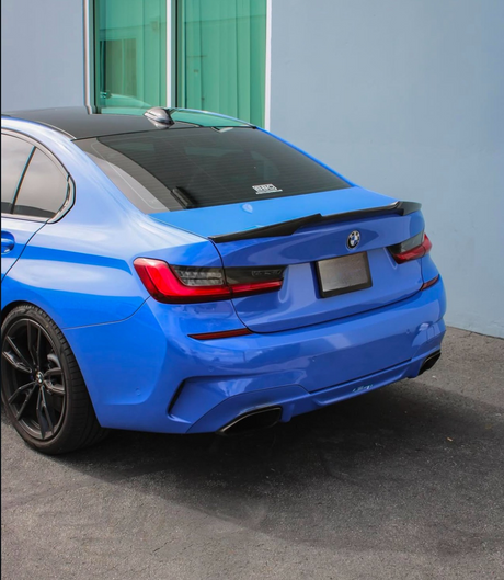 A blue car is parked near a light blue building. The car's rear features dual exhausts and dark-tinted windows. "BMW" and "3DDesign" are visible on the rear window.