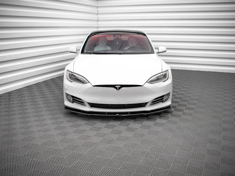 A white car, viewed from the front, is parked in a room with textured, gray-patterned flooring and walls featuring horizontal white panels.
