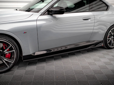 A sleek, silver car is parked on a textured gray floor. It features red brake calipers, sporty black wheels, and a side emblem with colored stripes.