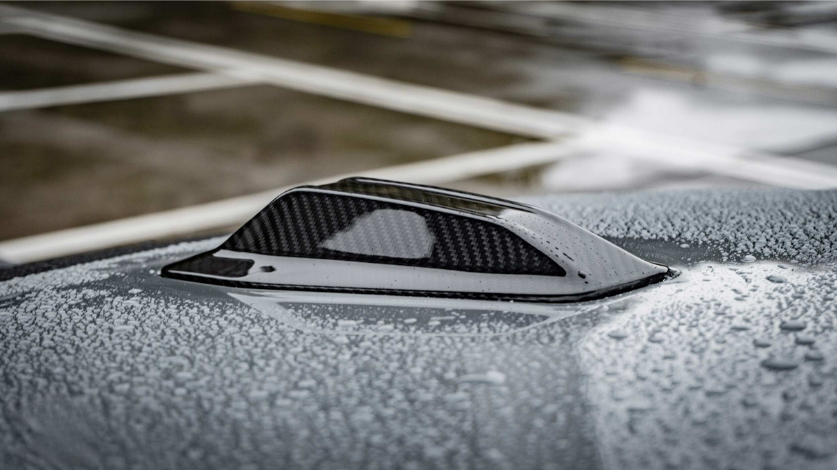 A carbon-fiber shark fin antenna rests on a car roof, covered with water droplets, in a parking lot with painted lines visible in the background.