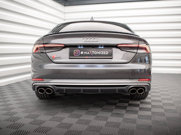 A gray Audi S5 is parked in a brightly lit showroom with white striped walls and a textured floor. The license plate reads "#MAXTONIZED" with an Instagram logo.