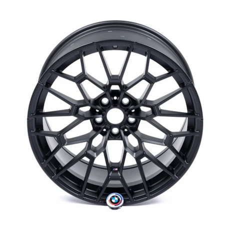 A black, intricately designed car wheel rim, featuring a multi-spoke pattern, is positioned in isolation on a white background. Near the bottom, a circular emblem displays a red and blue "M".