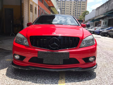 A red Mercedes car is parked on a sunlit street, surrounded by other vehicles and buildings. The front license plate area appears blurred, and the car's grille features the iconic logo.