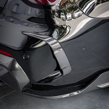 A sleek, glossy car bumper with a carbon fiber fin detail, reflecting nearby buildings, parked on a tiled surface.