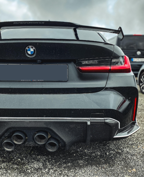 A black BMW car features quad exhaust pipes and a spoiler with "M Performance" text. It is parked on gravel beside another vehicle, under a cloudy sky.