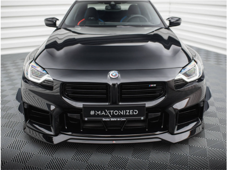 A shiny black BMW car, parked on a patterned surface, features a prominent front grill and headlights. The license plate reads "#MAXTONIZED Dealer BMW M-Cars." The background is a white, corrugated wall.