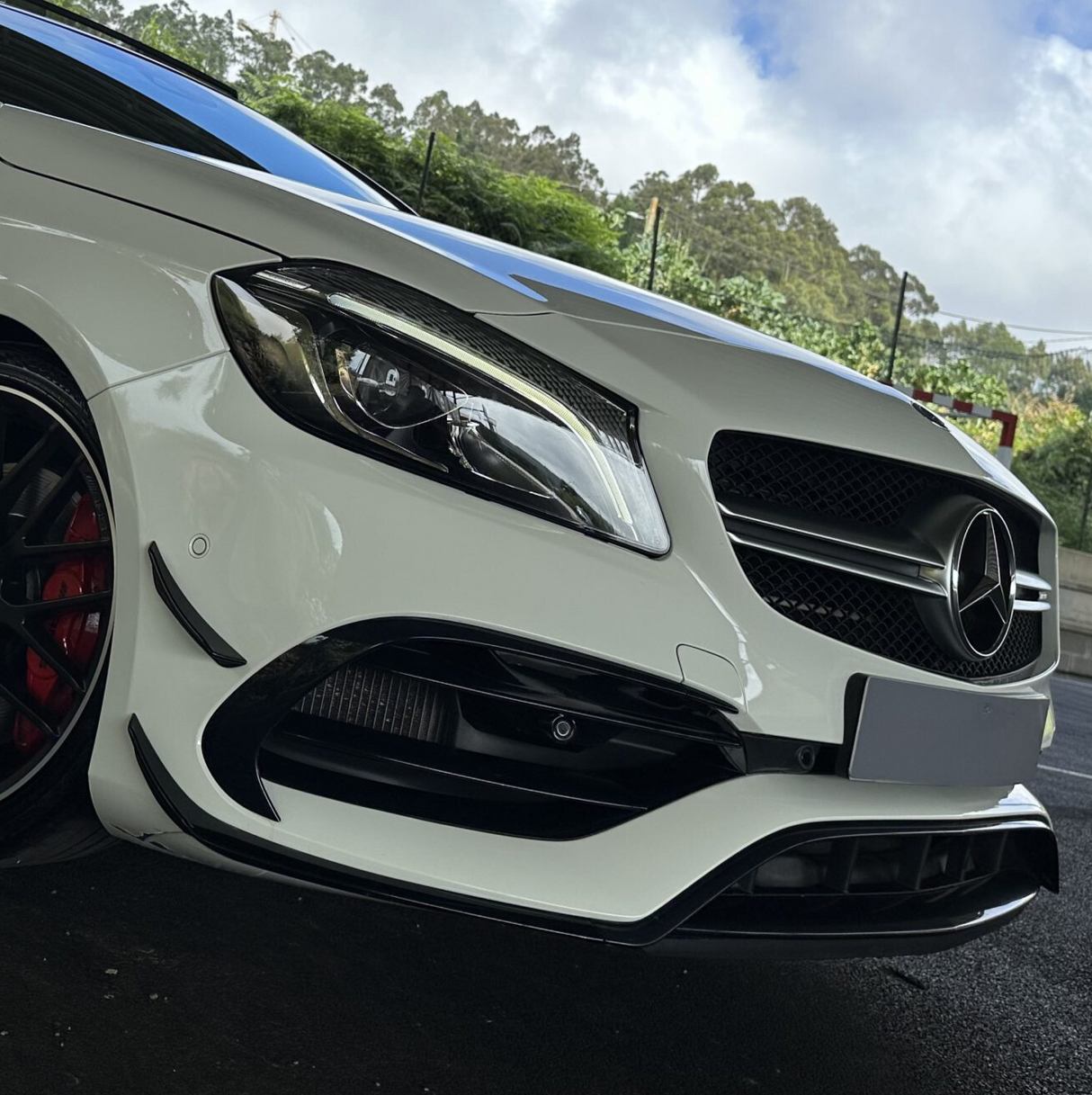 A white car with a prominent front grille and emblem is parked outdoors. The vehicle features sleek headlights and red brake calipers, set against a backdrop of greenery and a cloudy sky.