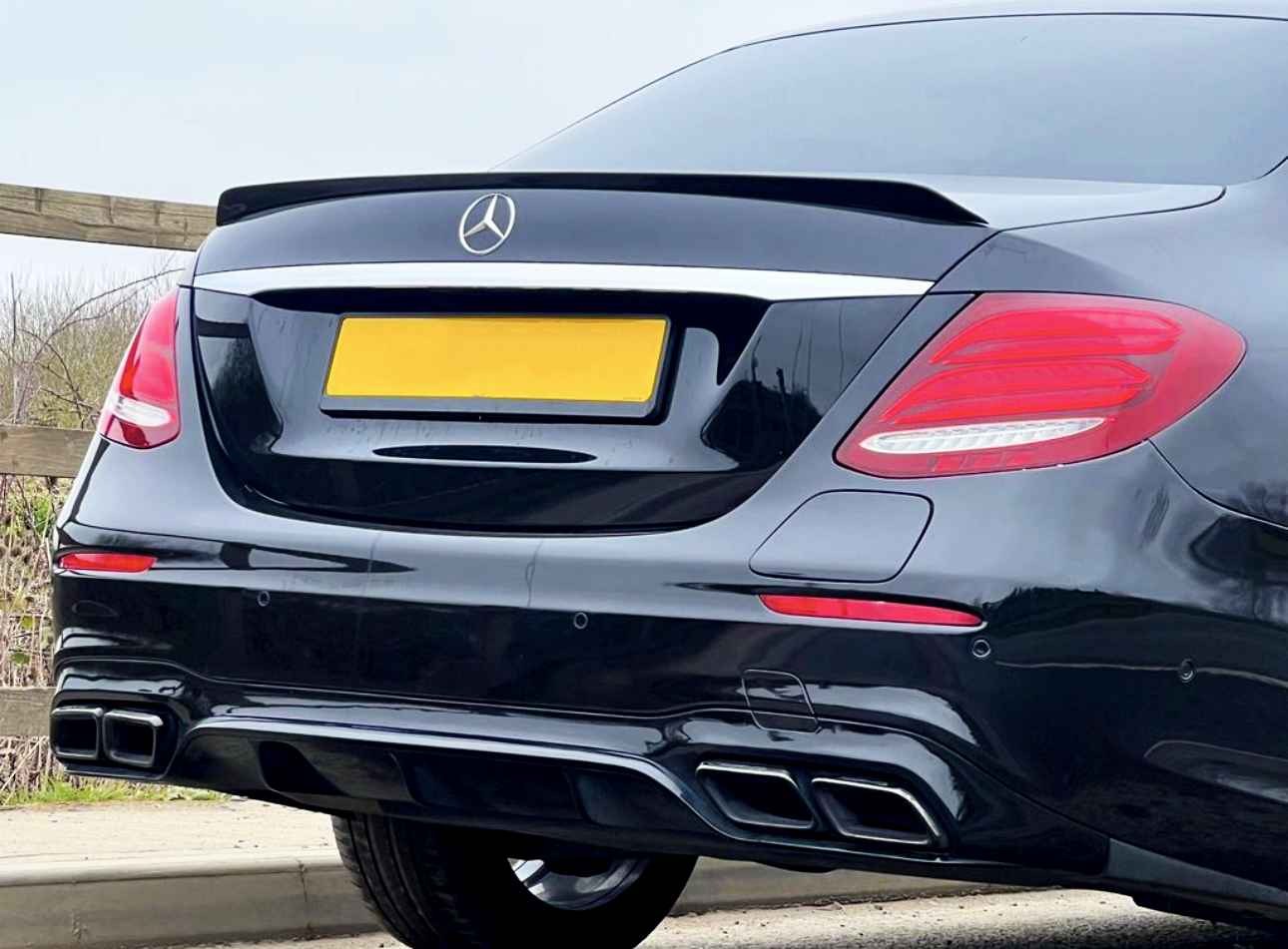 A black car with a visible rear, featuring Mercedes emblem, tinted windows, and dual exhausts, is parked on a road near wooden fencing and greenery. The license plate is yellow.