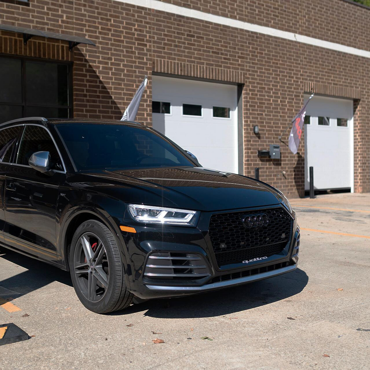 A black Audi SUV is parked in front of a brick building with white garage doors. The setting is a sunlit, concrete driveway, and the car's headlights reflect light. "Quattro" is visible on the grille.