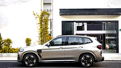 A silver SUV is parked beside a modern building with a black entrance. Yellow-green plants line the sidewalk. Reflections show other vehicles and signage: "INSTITUTO DE DRY CARBON".