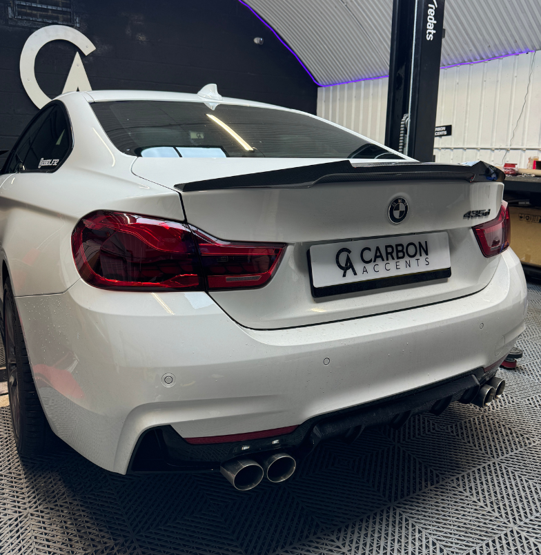 A white BMW 435d featuring a carbon fiber spoiler is parked inside a garage with a sign reading "CARBON ACCENTS." The garage has a dark back wall and a ribbed ceiling.